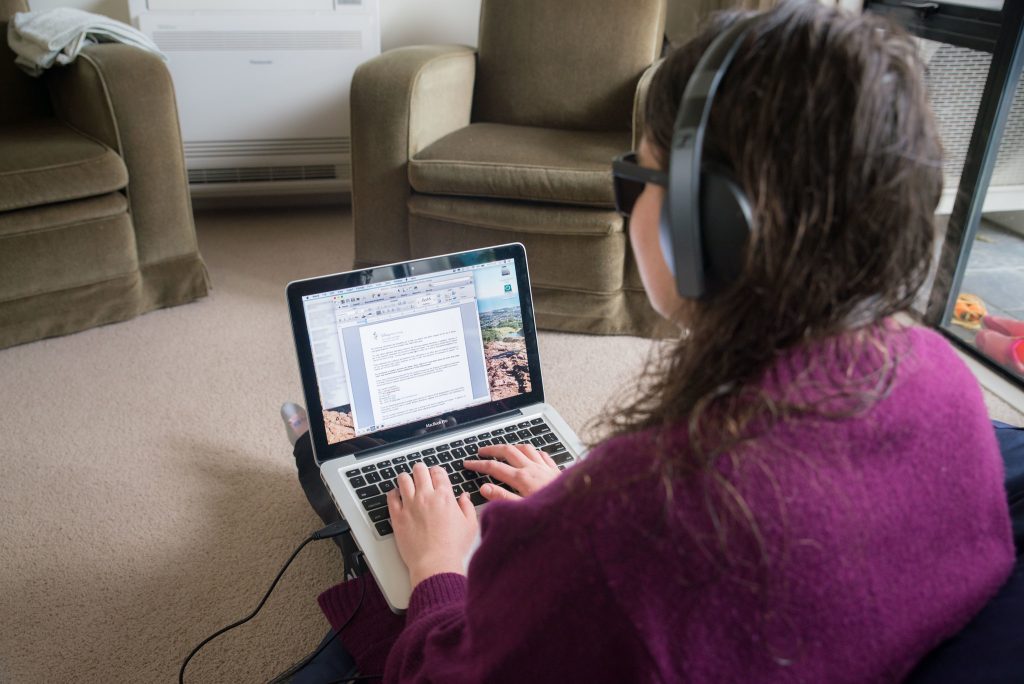 A person wearing headphones and a purple sweater is sitting on the floor and typing on a MacBook laptop