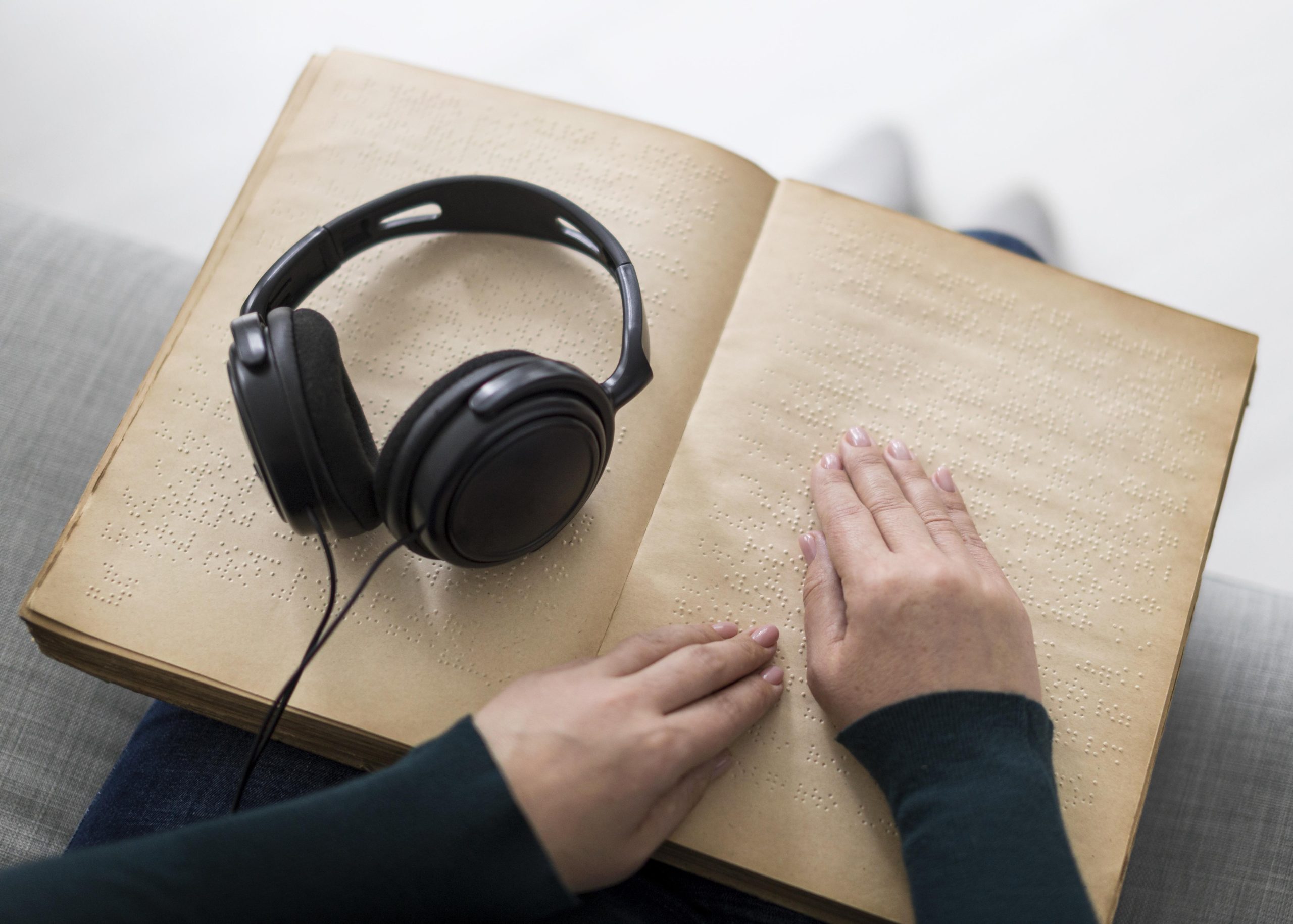 A person is reading a braille book using their hands, gently gliding fingers across the embossed dots. A pair of black over-ear headphones rests on the left page of the book