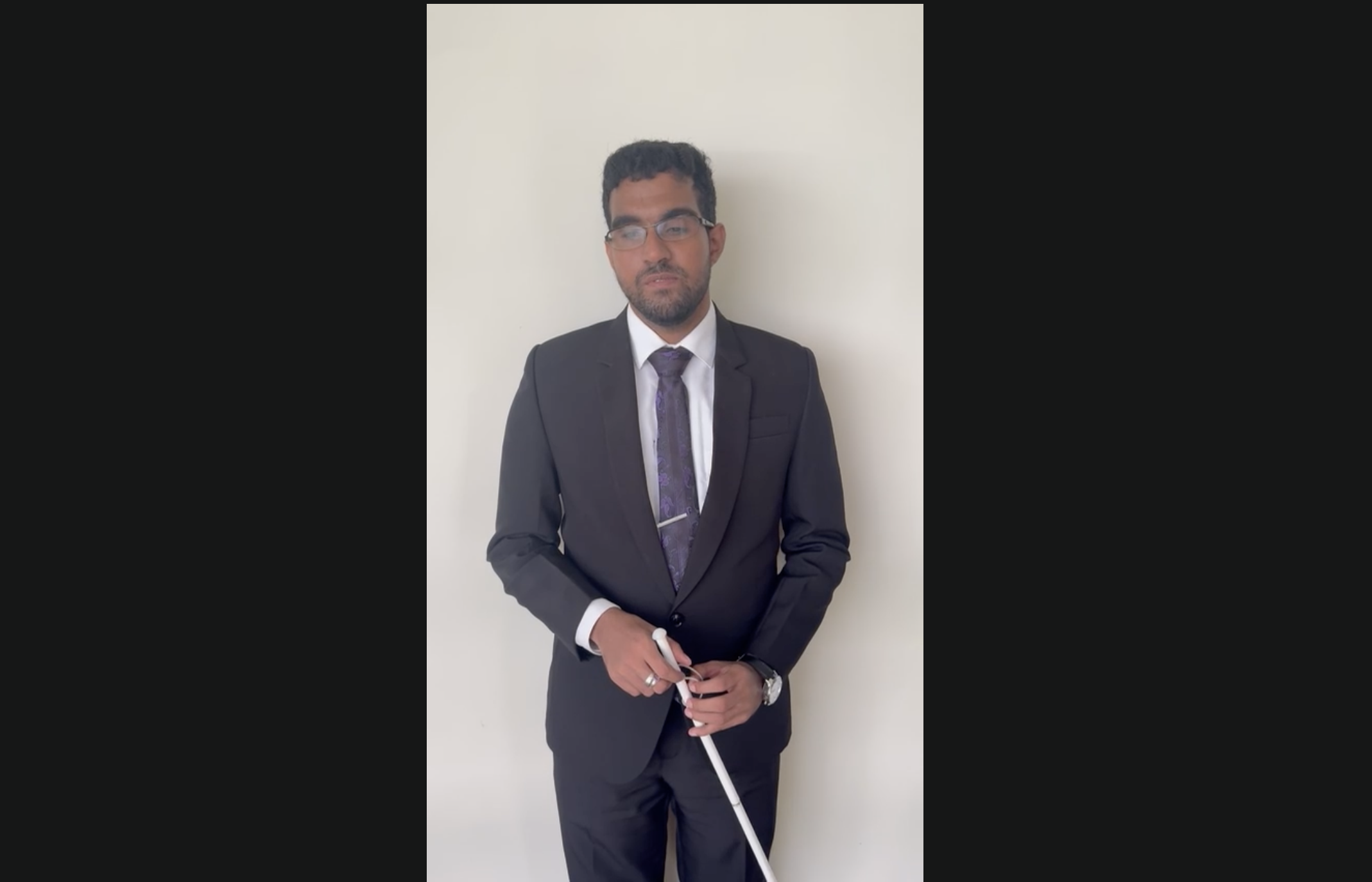 A medium shot of a man with glasses and a beard, formally dressed in a black suit and tie, standing in front of a plain wall while holding a long white cane.